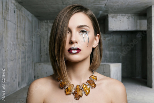 Futuristic cyberpunk woman portrait with liquid silver metallic tear makeup and amber necklace posing in concrete brutalist architecture industrial room