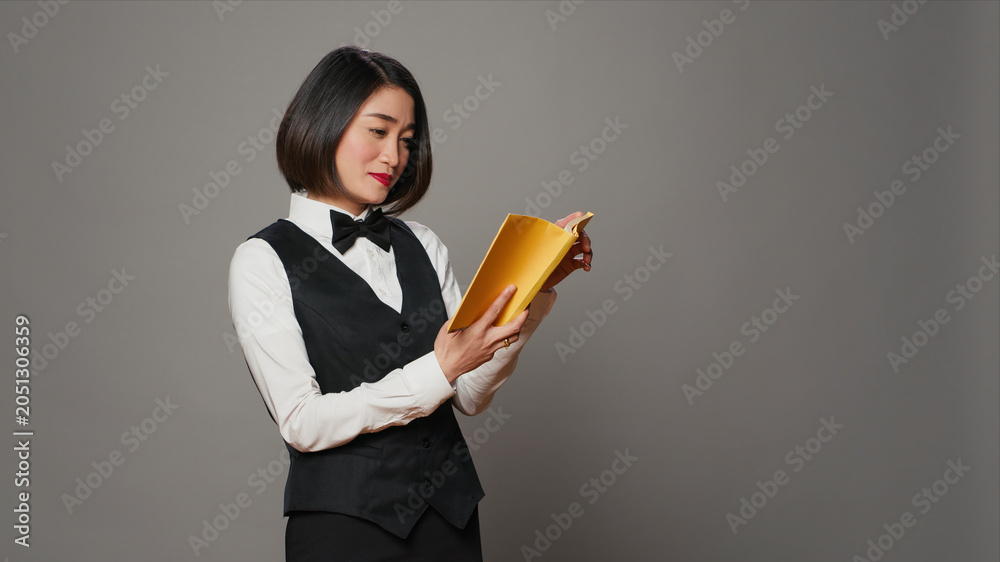 Obraz premium Asian receptionist reading a literature book over grey background, enjoying lecture hobby with a novel story in studio, hotel concierge employee reads a fiction tale for knowledge