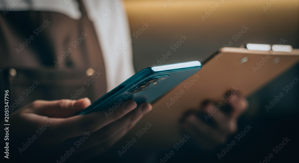 Fototapeta premium Worker in an apron holding a smartphone and clipboard representing digital business management and workflow efficiency.