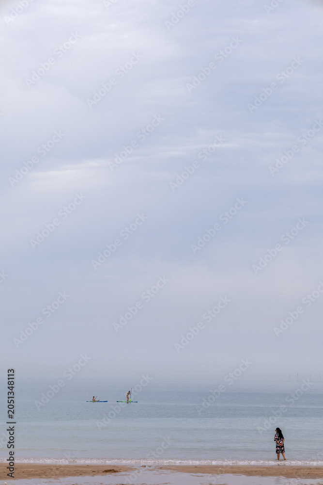 Fototapeta premium Calm beach with lone woman and distant paddle boarders.Calm beach scene with a lone woman at the shoreline and distant paddle boarders on a misty sea under a soft cloudy sky.