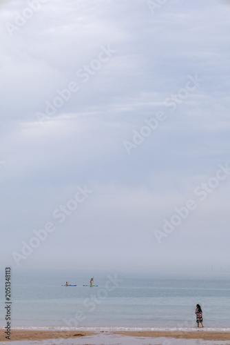 Calm beach with lone woman and distant paddle boarders.Calm beach scene with a lone woman at the shoreline and distant paddle boarders on a misty sea under a soft cloudy sky.