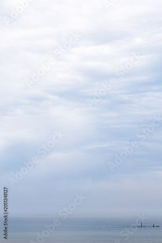 Calm sea and cloudy sky with distant paddle boarders.Calm beach scene with two people paddle boarding on a still sea under a soft cloudy sky. Peaceful coastal view with gentle waves, wet sand, and lot