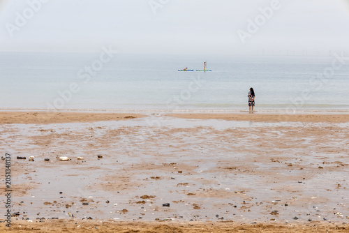 Woman standing on quiet beach at low tide.Calm beach scene with two people paddle boarding on a still sea under a soft cloudy sky. Peaceful coastal view with gentle waves, wet sand, and lots of open c