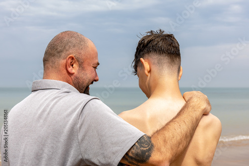 Father and teenage son sharing a quiet beach moment.Father with arm around his teenage son standing on a beach, seen from behind, sharing a quiet supportive moment by the sea under a cloudy sky.