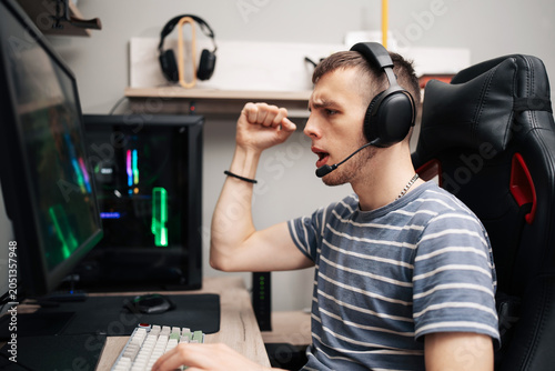 An excited young gamer wearing a headset celebrates a victory during an intensive online multiplayer video game session at his desktop computer setup.