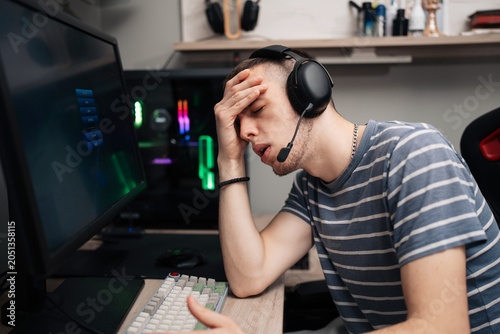 A young male gamer wearing a headset expresses frustration and fatigue, holding his head in disappointment while playing an intense online multiplayer game at his computer desk.