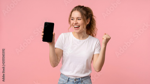 young cute woman in white t-shirt shows blank smartphone screen and rejoices in victory on pink isolated background, the winner girl advertises copy space on the phone, mockup