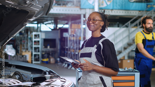 Фотография Portrait of smiling engineer doing car maintenance in repair shop using tablet to keep track of operations, with qualified specialist in garage looking over checklist on device while fixing vehicle