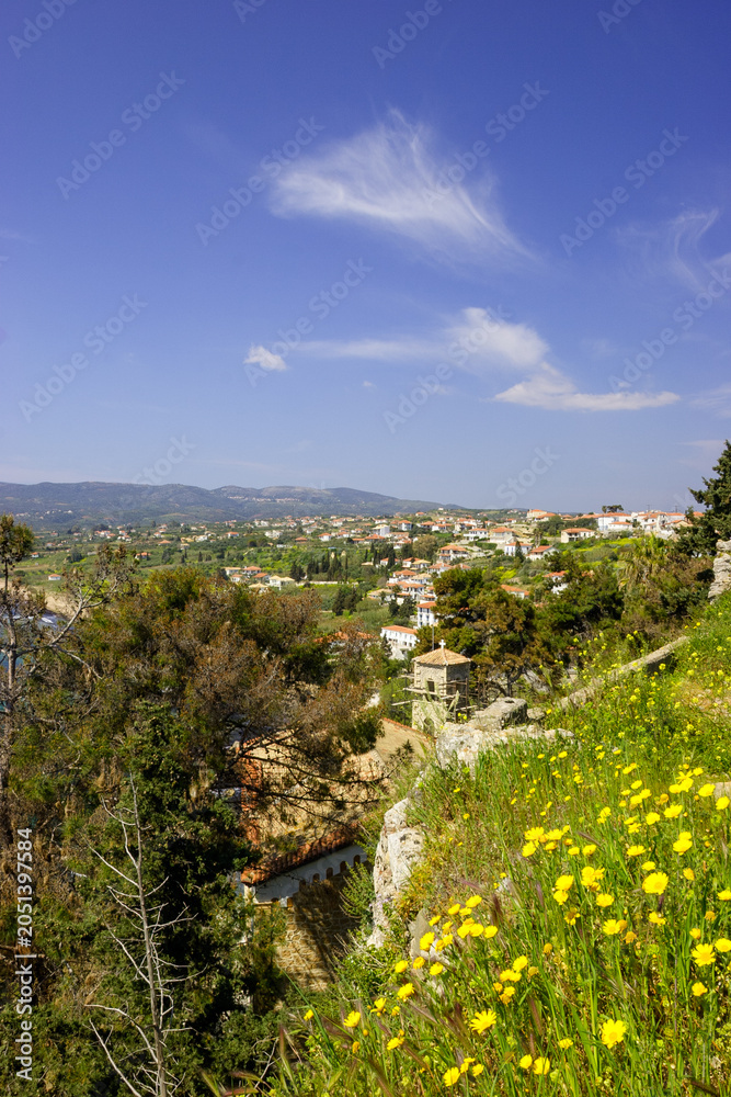 Obraz premium A scenic view of a coastal town with red-tiled roofs and white buildings, nestled among green hills under a bright blue sky with wispy clouds. 