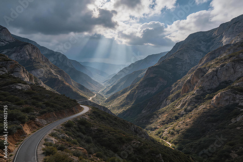 Winding Mountain Road Through Scenic Valley Under Dramatic Cloudy Sky with Sun Rays