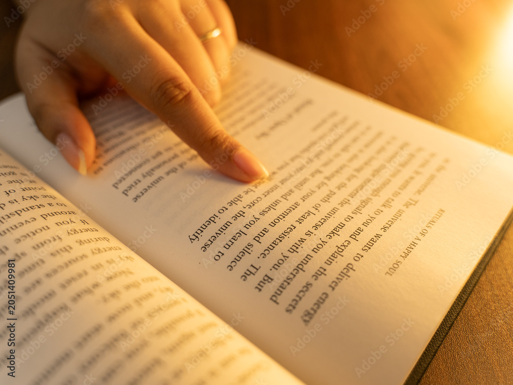 Obraz premium Close-up of a woman's hands holding an open book with clearly visible text pages, shot in warm cozy indoor lighting with a soft glowing table lamp in the background, evoking reading and knowledge. 