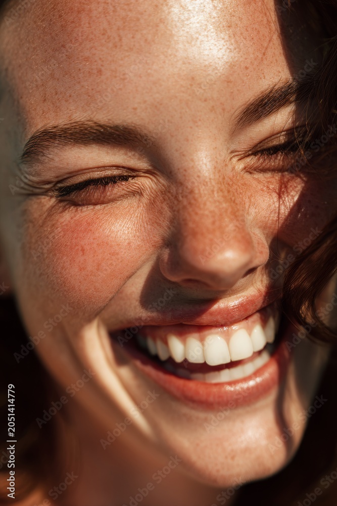 Fototapeta premium Close up portrait of a joyful young woman with freckles laughing with closed eyes in bright warm sunlight