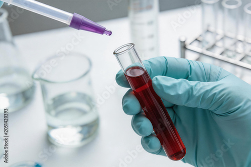 Gloved hand holding test tube with red liquid and dropper in laboratory setting