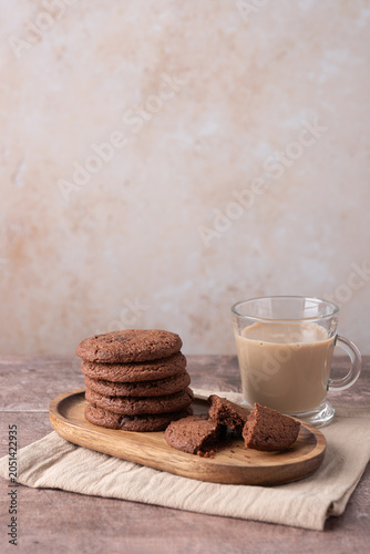 Broken chocolate cookies on a wooden tray with milk coffee in a glass cup, horizontal scene on wooden table and beige stone background with copy space