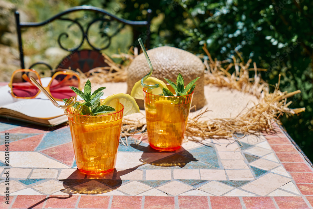 Obraz premium Summer refreshing lemonade drink or non alcoholic cocktail with lemon slices in orange drinking glasses on the table in the garden. Fresh healthy cold lemon beverage. Water with lemon. Summer mood.