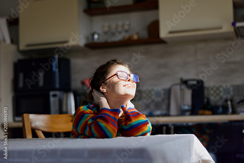 Photography A thoughtful girl in a colorful striped sweater sits at a table in a warm home kitchen, gazing upward with a hopeful smile, conveying curiosity and calm