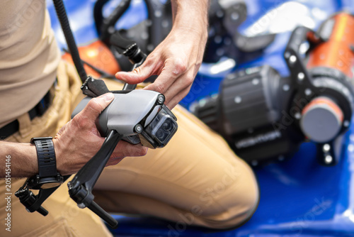 Male technician in beige shirt inspects small drone device while seated on blue tarp, with additional equipment visible in the background