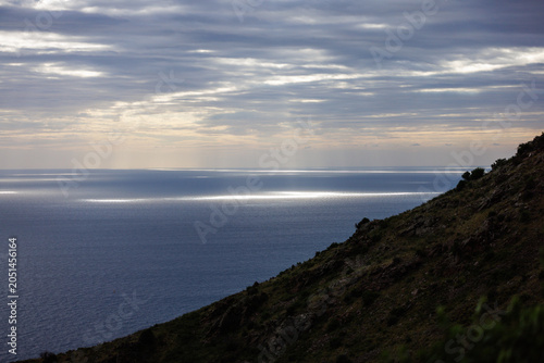 Paysage côtier à l'aube avec une mer calme et des rayons de soleil perçant les nuages. Une colline rocheuse et légèrement végétalisée domine le premier plan.