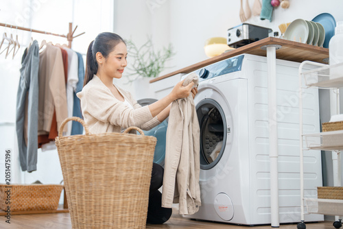 Laundry woman washing machine basket clothing home chore clean fabric room, smiling woman loading clothing into washing machine beside wicker basket in bright laundry room with hanging garment