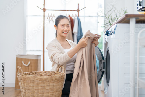 Laundry woman washing machine basket clothing home clean housework smile fabric young Asian woman loading beige clothing into washing machine beside wicker basket in bright home laundry room