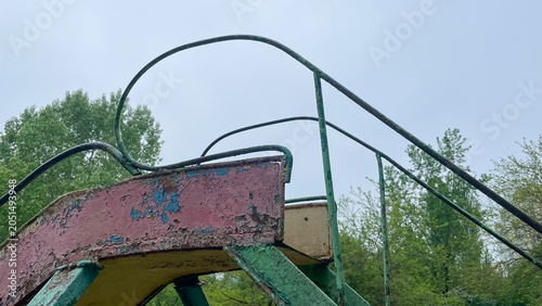 Old rusty metal playground slide with peeling paint against green trees