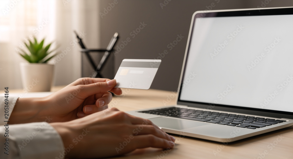 Fototapeta premium Hands holding a credit card near a laptop. Person making an online payment at a desk. E-commerce and digital banking concept