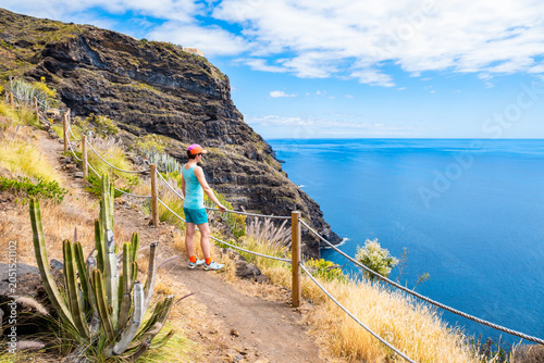 Woman in hiking attire stands on a dirt path with rope railings, looking out at a massive sea cliff at Poris de la Candelaria, La Palma, Canary Islands, Spain