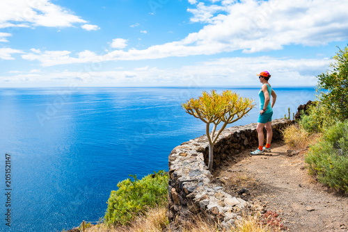 Hiker stands on a stone path next to a low rock wall, looking over a wide expanse of blue ocean at Poris de la Candelaria, La Palma, Canary Islands, Spain