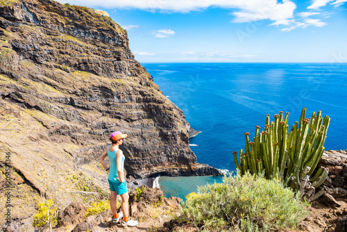 Hiker in a bright cap stands by a large cardon cactus, admiring a hidden beach and massive sea cliffs at Poris de la Candelaria, La Palma, Canary Islands, Spain