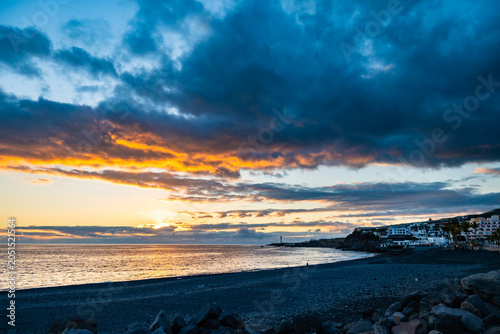 Dramatic sunset with fiery orange clouds over a dark volcanic sand beach and coastal Puerto Naos town, La Palma, Canary Islands, Spain