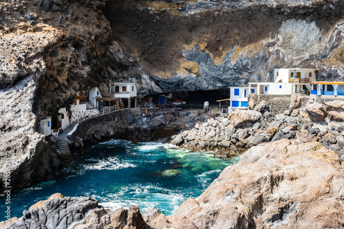 Quaint white houses with blue accents are nestled directly inside a massive volcanic cave over a rocky cove at Poris de la Candelaria, La Palma, Canary Islands, Spain