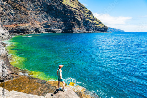 Hiker in a pink cap looks out over a vibrant turquoise ocean bay next to towering volcanic cliffs at Puntagorda, La Palma, Canary Islands, Spain