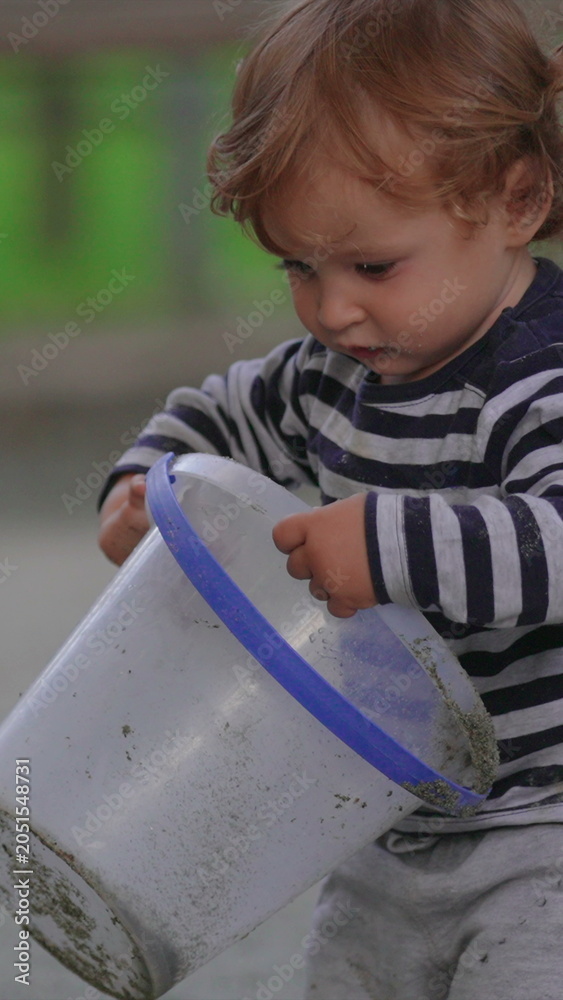 Fototapeta premium Young child examining colorful toy bucket at public playground during outdoor activity highlighting curiosity, sensory discovery and playful early childhood learning