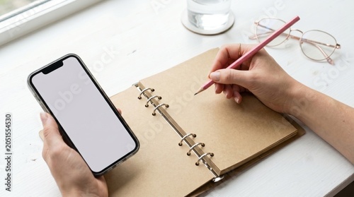 Woman using smartphone and writing in notebook on white desk with glasses and water glass