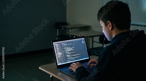 Over the shoulder shot of a student programming on laptop in a dark classroom with blank blackboard copy space