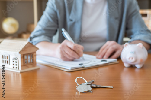 Person signing crucial documents for a mortgage loan, buying a new house, or making a real estate investment, with a piggy bank and house keys on the desk symbolizing saving and ownership