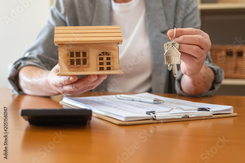 Real estate agent at desk holding wooden house model and keys over mortgage documents with pen and calculator, symbolizing home purchase, financing and property investment planning
