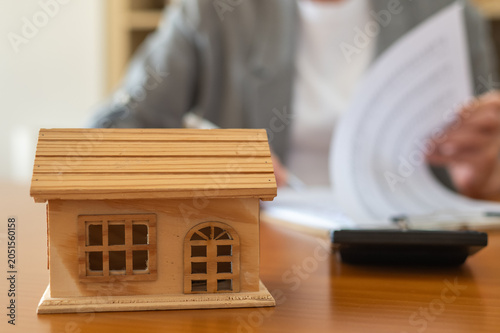 Small wooden house model on a desk with blurred person reviewing documents and calculator, symbolizing mortgage, home purchase contracts, property financing and real estate investment decisions