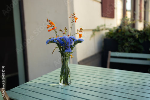Close-up shot of a lovely bouquet with blue field flowers standing in a simple glass jar on a green wooden table at an outdoor cafe terrace on a sunny day.