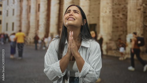 Fényképezés Young woman praying with clasped hands at a historic stone building facade, looking upward among pedestrians in a plaza; plea hope