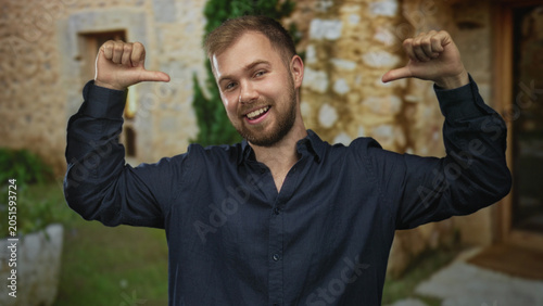 Slika na platnu Man smiling and points thumbs to chest at building entrance outdoors, wearing navy shirt and beard visible; confidence