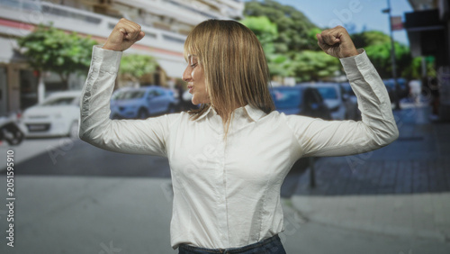 Photos Woman flexes both arms with clenched fists on a busy street, smiling and turning her head to the side while wearing a white shirt and jeans; confidence empowerment