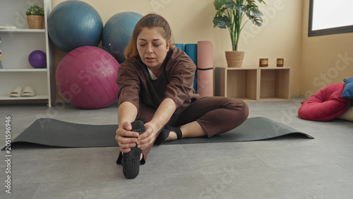 Bild auf Leinwand Woman seated on a yoga mat reaches forward and grasps her foot with both hands, stretching hamstring and ankle in a studio; serenity mindfulness
