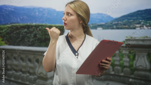 Quadro su tela Woman holding pink clipboard points thumb behind and speaks on a building terrace by a lake with mountains in view; surprise instruction