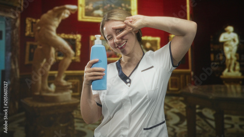 Slika na platnu Woman nurse holding blue pump bottle, smiling with eyes closed and making peace sign while wearing uniform in building; cheerfulness care