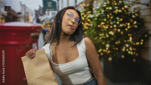 Quadro su tela Woman holding paper bag and sticking tongue out on street with christmas tree and red postbox; playful defiance
