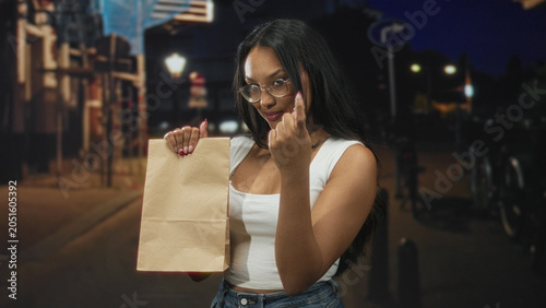 Bilde på lerret Young hispanic brunette woman wearing glasses holds a paper bag and beckons with hand on a dim street at night; confidence invitation