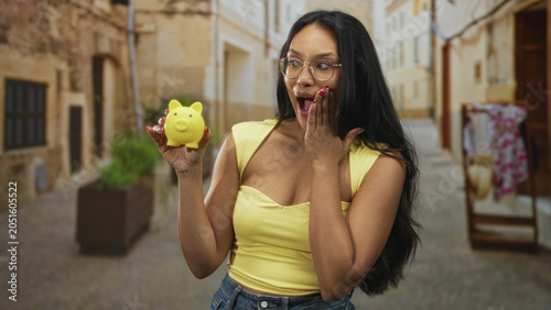 Konstfotografi Woman holding yellow piggybank with hand on chest, wearing glasses and yellow top on street; surprise saving optimism