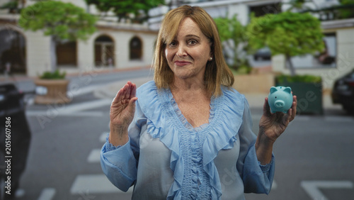 Photos Woman holding turquoise piggy bank in right hand and pinching fingers gesture on a street; saving prudence