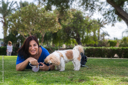 A smiling woman with her Shih Tzu lying on the grass. Outdoor pet lifestyle. She smiles affectionately at her dog. A small, brown and white dog sniffs her hand.